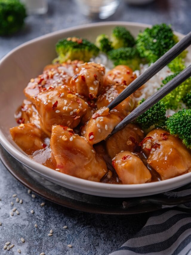 A plate of orange chicken garnished with sesame seeds and a pair of chopsticks is grabbing a piece of chicken.