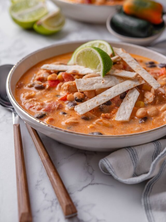 A bowl of creamy soup with diced tomatoes, corn, beans, and chicken, with crispy tortilla strips and lime wedges garnished on top.