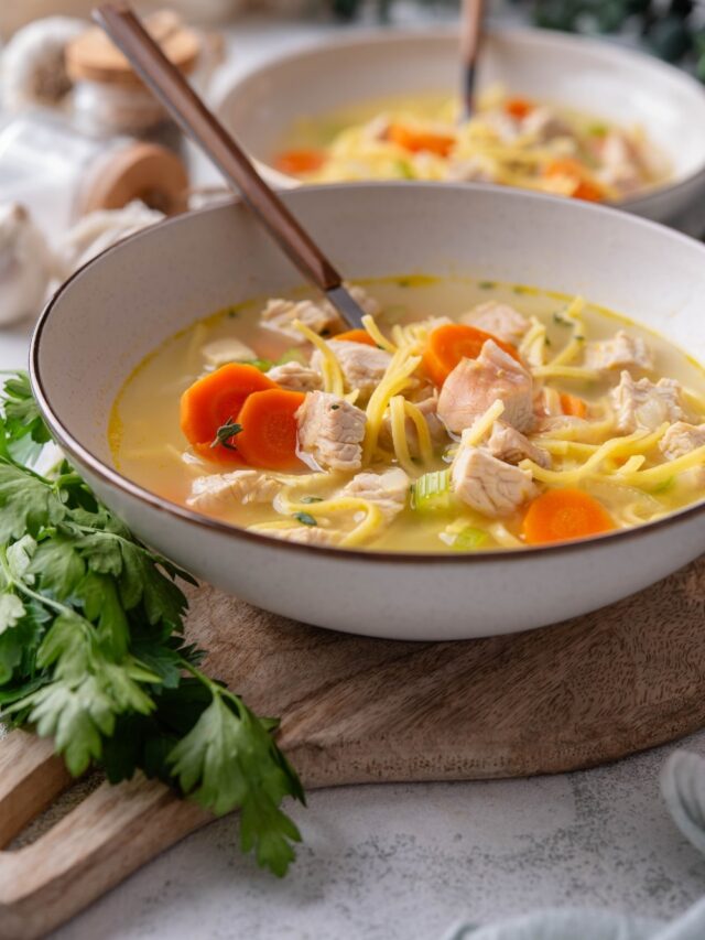A bowl of turkey noodle soup with chunks of carrots and celery. There is a spoon in the bowl and a second bowl in the background.