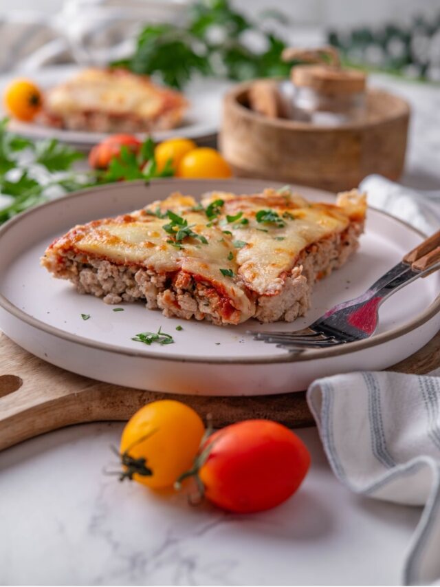Two slices of crustless pizza garnished with fresh parsley on a white ceramic plate. The plate is on a wooden serving board and has two forks. There is a second plate of crustless pizza and a wooden bowl in the background.