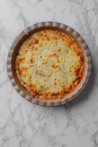 A pie plate with cooked crustless pizza and melted cheese on top. The pie plate is on a marble countertop.