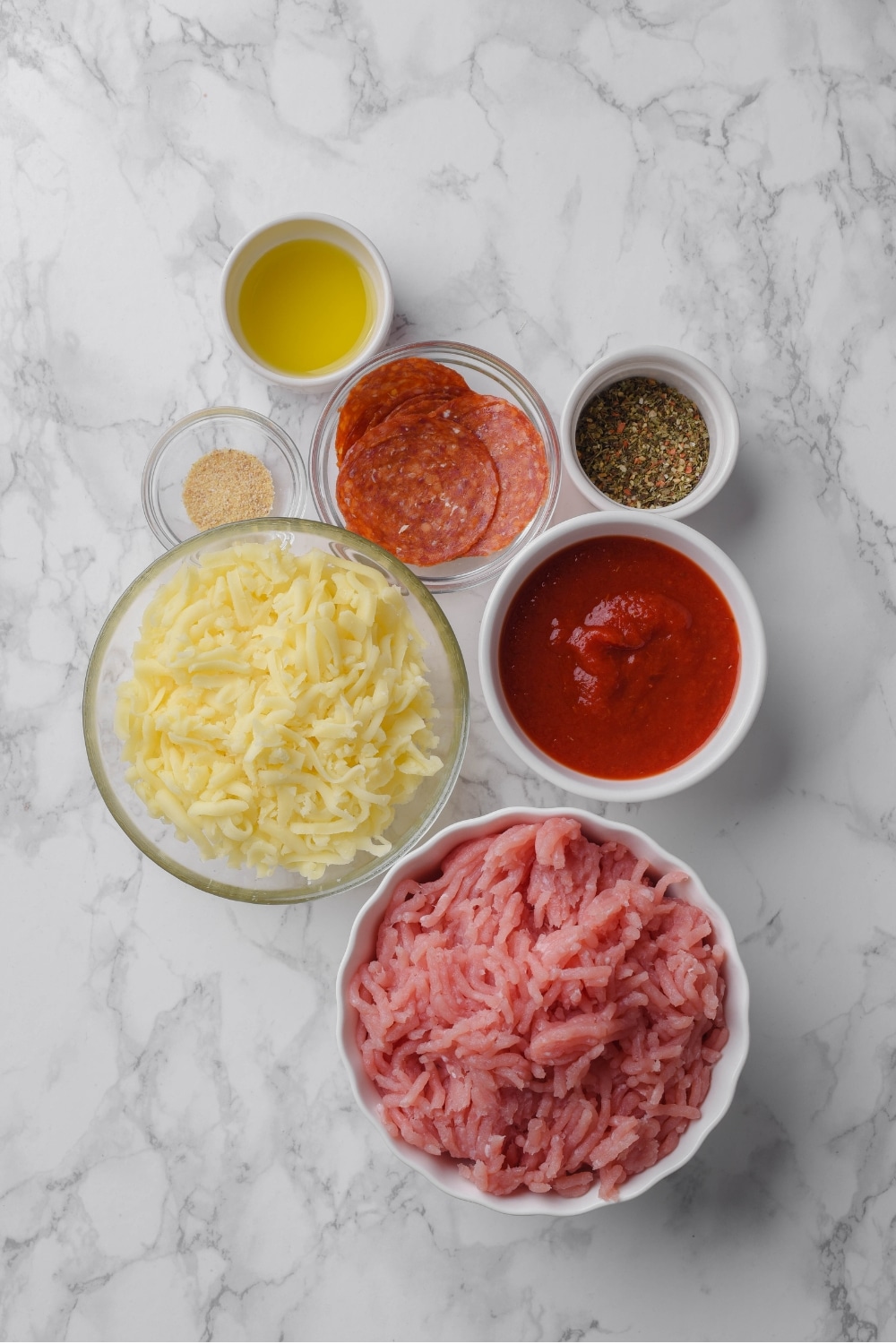 An assortment of glass and white ceramic bowls filled with shredded cheese, raw ground turkey, pizza sauce, sliced pepperoni, italian seasoning, garlic, and olive oil.