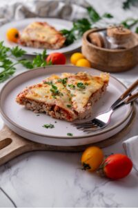 Two slices of crustless pizza garnished with fresh parsley on a white ceramic plate. The plate is on a wooden serving board and has two forks. There is a second plate of crustless pizza and a wooden bowl in the background.