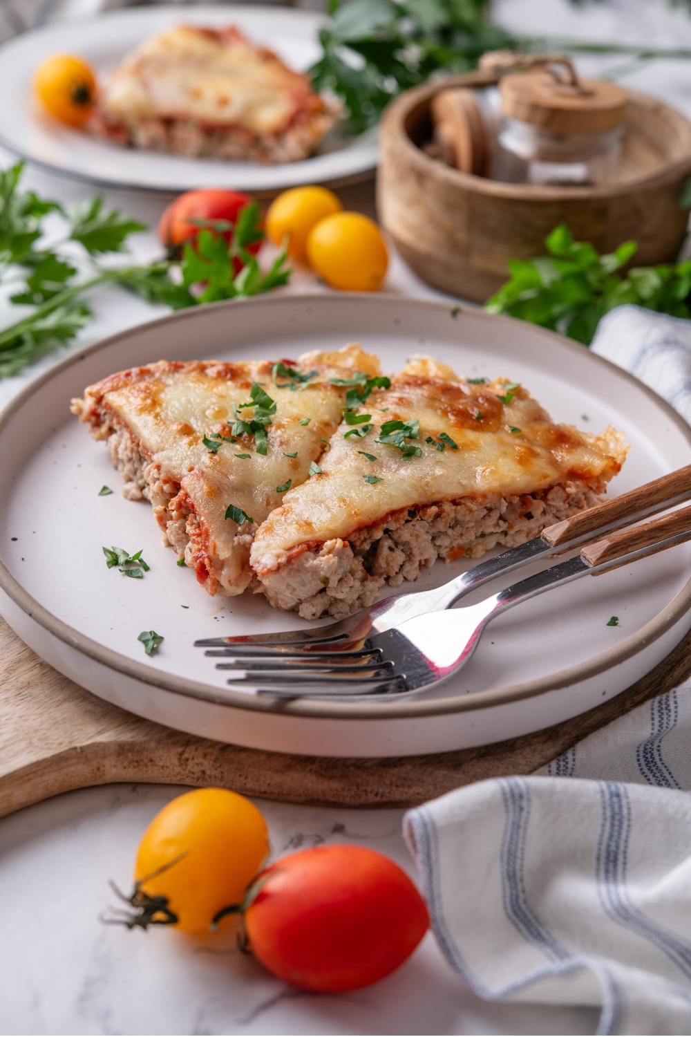 Two slices of crustless pizza garnished with fresh parsley on a white ceramic plate. The plate is on a wooden serving board and has two forks. There is a second plate of crustless pizza and a wooden bowl in the background.