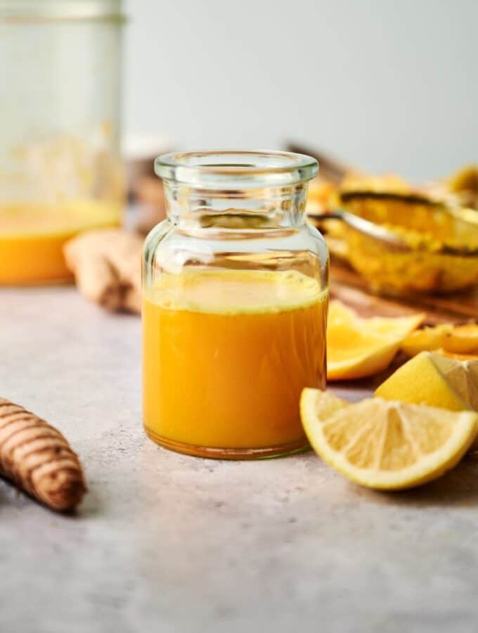 A lemon wedge in front of a large glass that has a ginger turmeric shot in it.