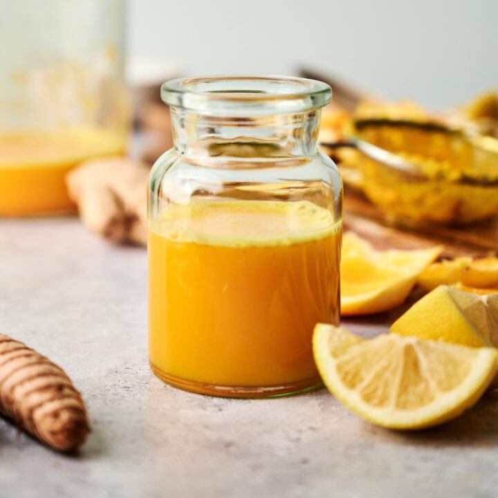 A lemon wedge in front of a large glass that has a ginger turmeric shot in it.