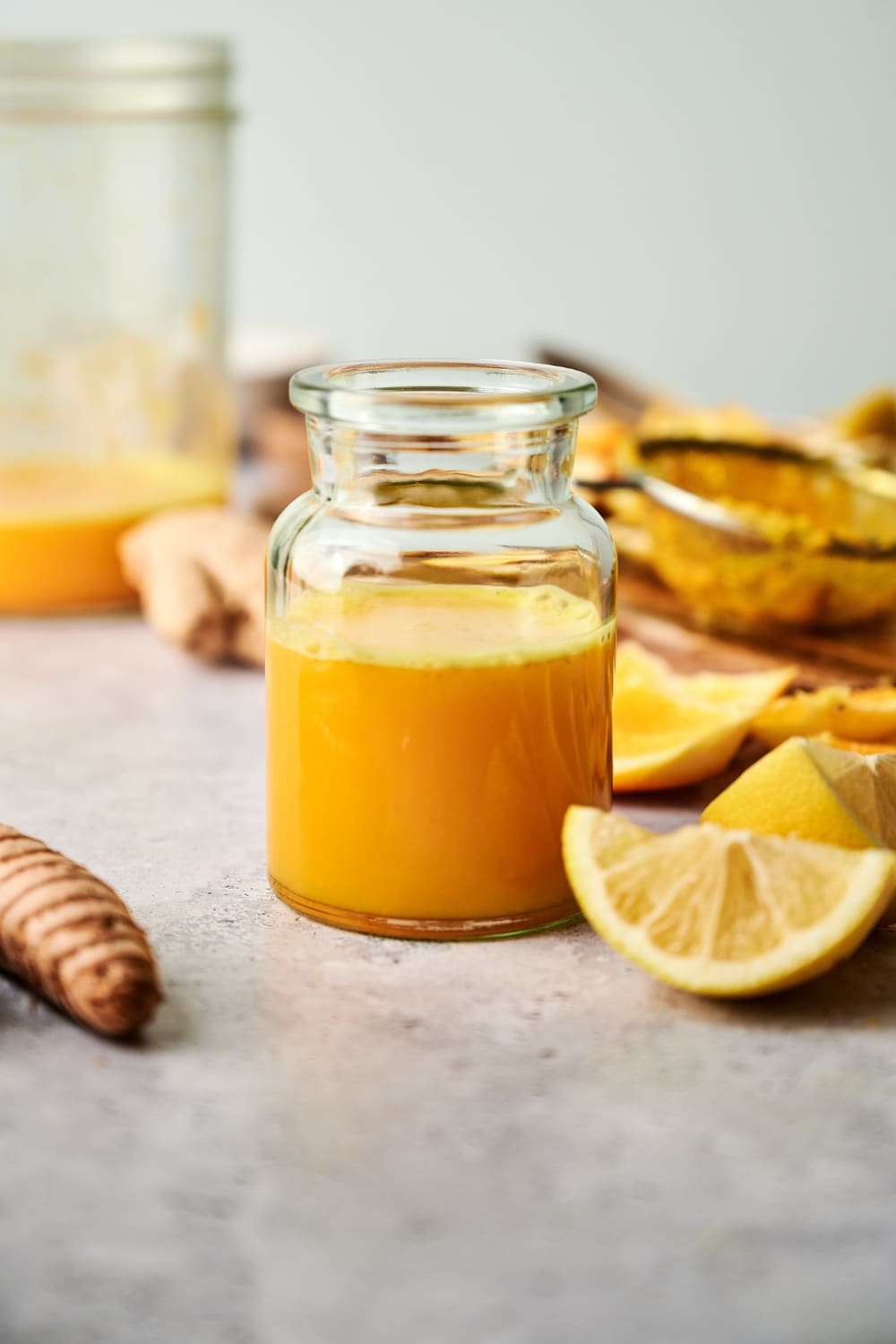 A lemon wedge in front of a large glass that has a ginger turmeric shot in it.