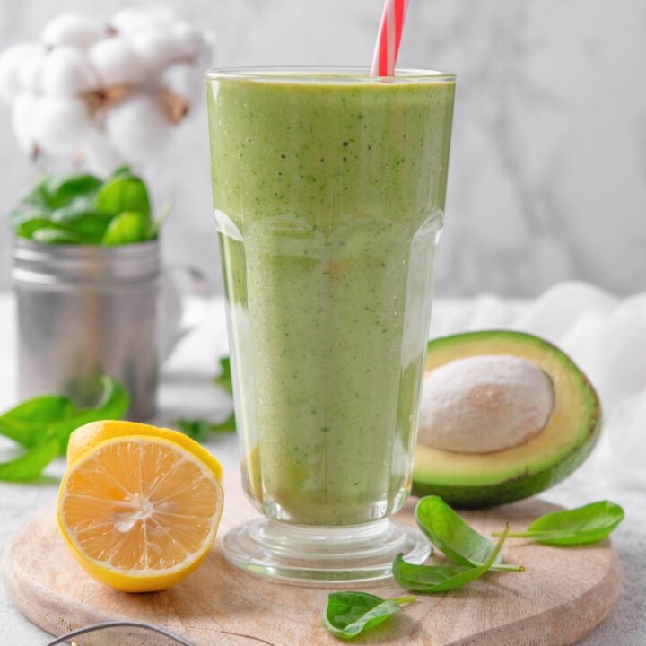 A glass cup that is filled with a green smoothie. A red and white striped straw is in the cup. The cup is on a wooden cutting board with half an avocado and orange on it.
