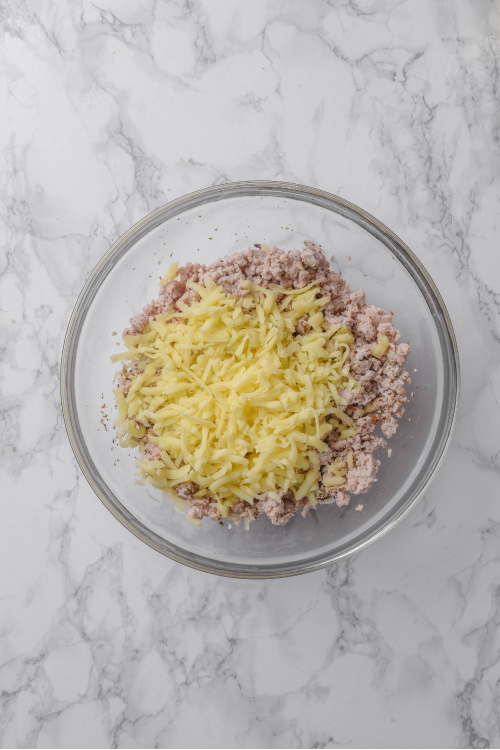 A glass bowl filled with cooked ground turkey and shredded cheese on top a white marble countertop.