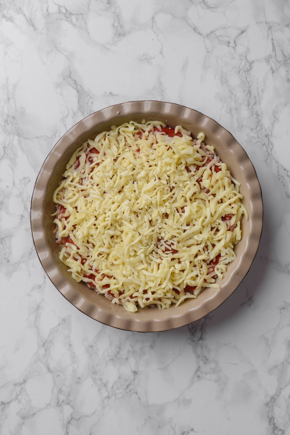 A pie pan filled with ground turkey layered with pizza sauce and cheese. The pie pan in on a marble countertop.