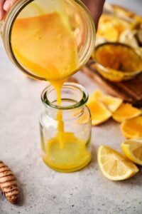 A hand holding a glass pouring a ginger turmeric shot into a small glass.
