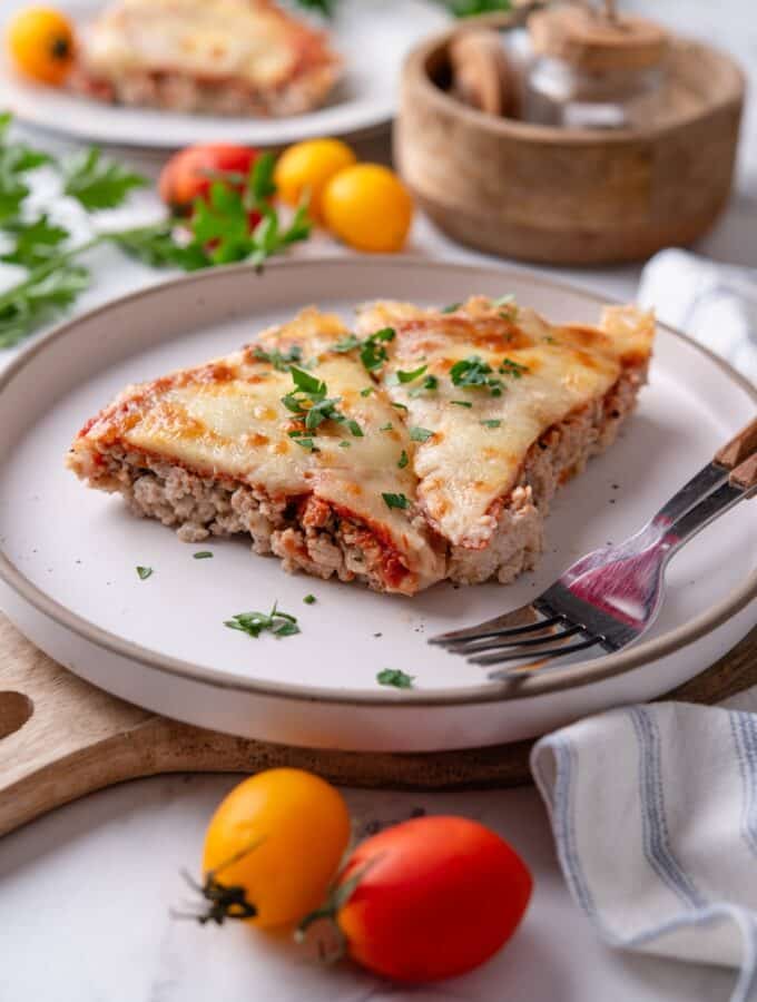 Two slices of crustless pizza garnished with fresh parsley on a white plate and two forks. The plate is on top of a serving board with a striped napkin. There is a second plate of crustless pizza and a wooden bowl in the background.