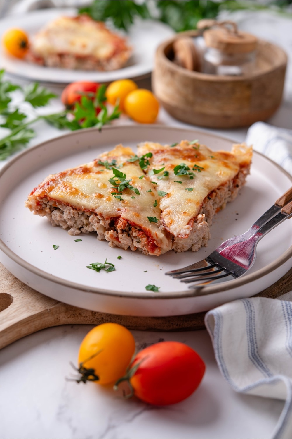 Two slices of crustless pizza garnished with fresh parsley on a white plate and two forks. The plate is on top of a serving board with a striped napkin. There is a second plate of crustless pizza and a wooden bowl in the background.