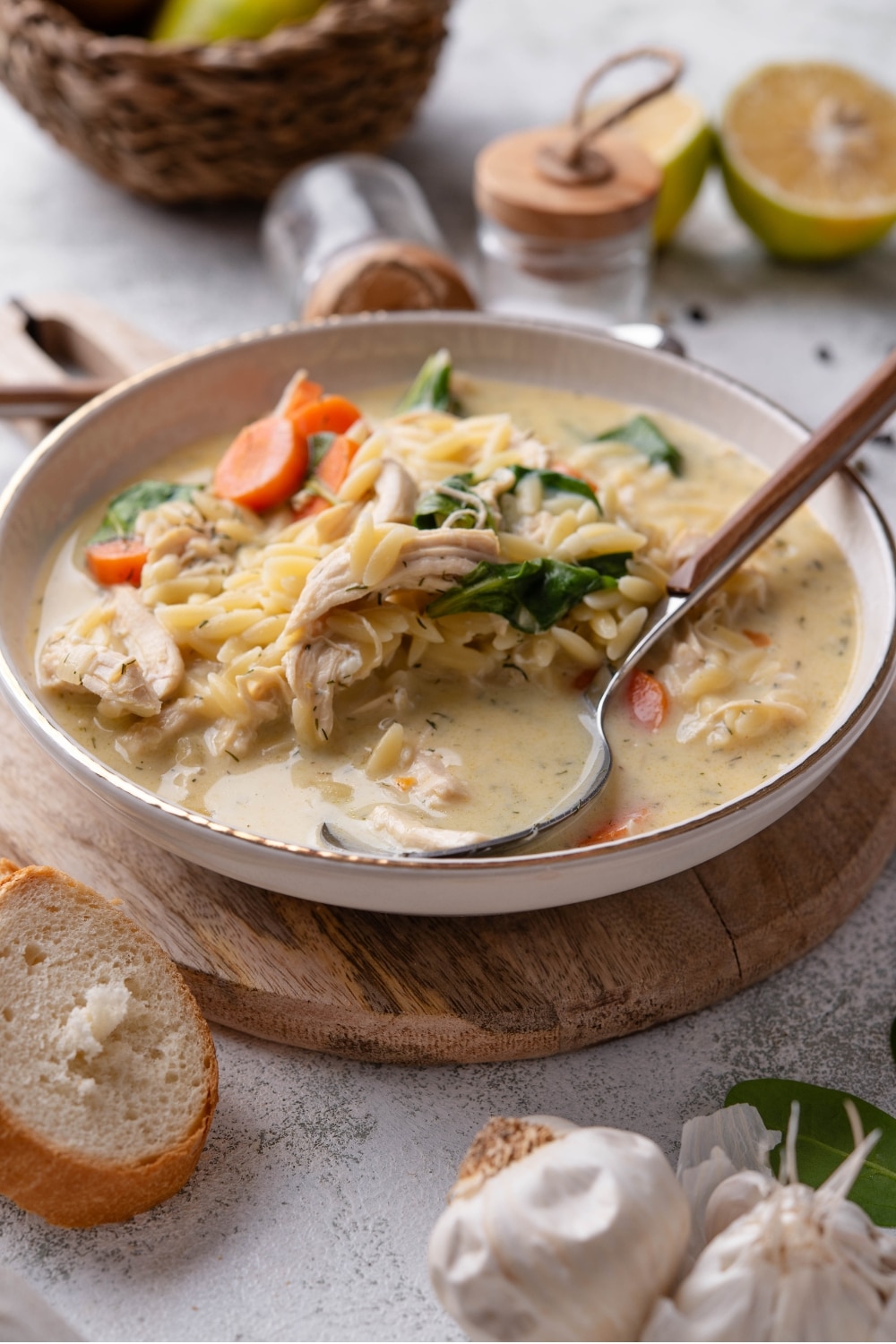 A white ceramic bowl of lemon chicken soup with orzo pasta, carrots, and spinach. The bowl sits on a wooden board with a spoon resting in the soup.