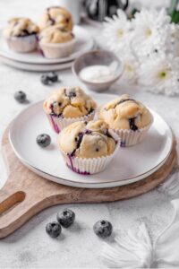 Three blueberry muffins sit on a white ceramic plate atop a wooden serving board. There is an extra plate of muffins and white flowers in the background
