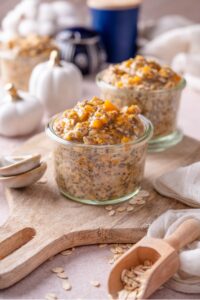 Two glass jars filled with pumpkin overnight oats on a wooden serving board. There are decorative white pumpkins and ceramic spoons next to them.