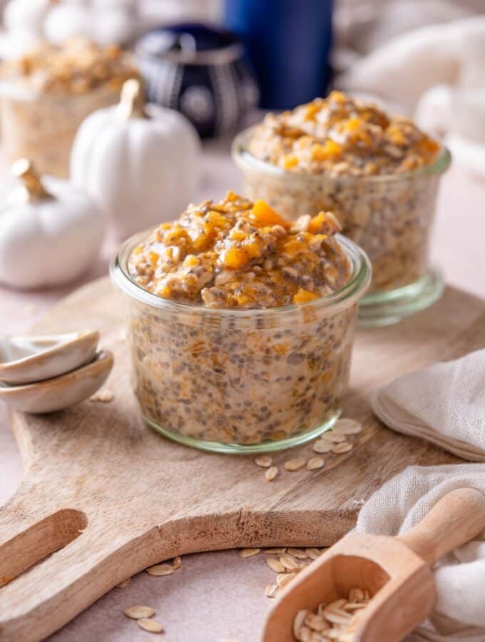 Two glass jars filled with pumpkin overnight oats on a wooden serving board. There are decorative white pumpkins and ceramic spoons next to them.