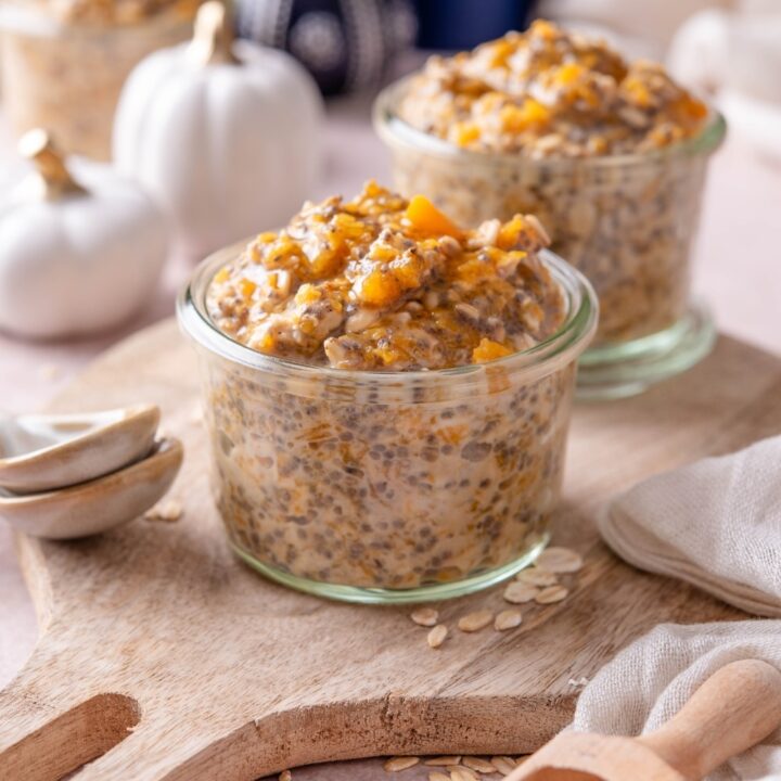 Two glass jars filled with pumpkin overnight oats on a wooden serving board. There are decorative white pumpkins and ceramic spoons next to them.