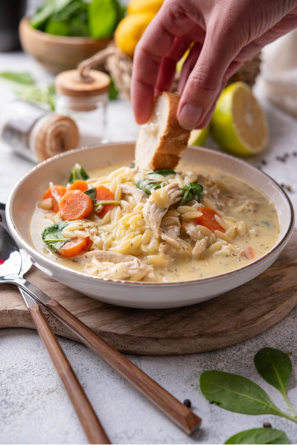 A white ceramic bowl of lemon chicken soup with orzo pasta, shredded chicken, carrots, and spinach. A hand is dipping a slice of bread into the soup.