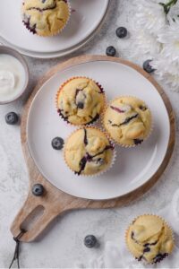 Three blueberry muffins displayed on a white ceramic plate sitting on a wooden serving board. The tray is on a marble counter with additional muffins next to it.