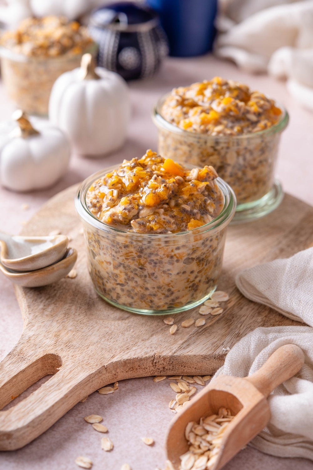 Two jars of pumpkin overnight oats on a wooden serving board with ceramic spoons and decorative pumpkins in the background.