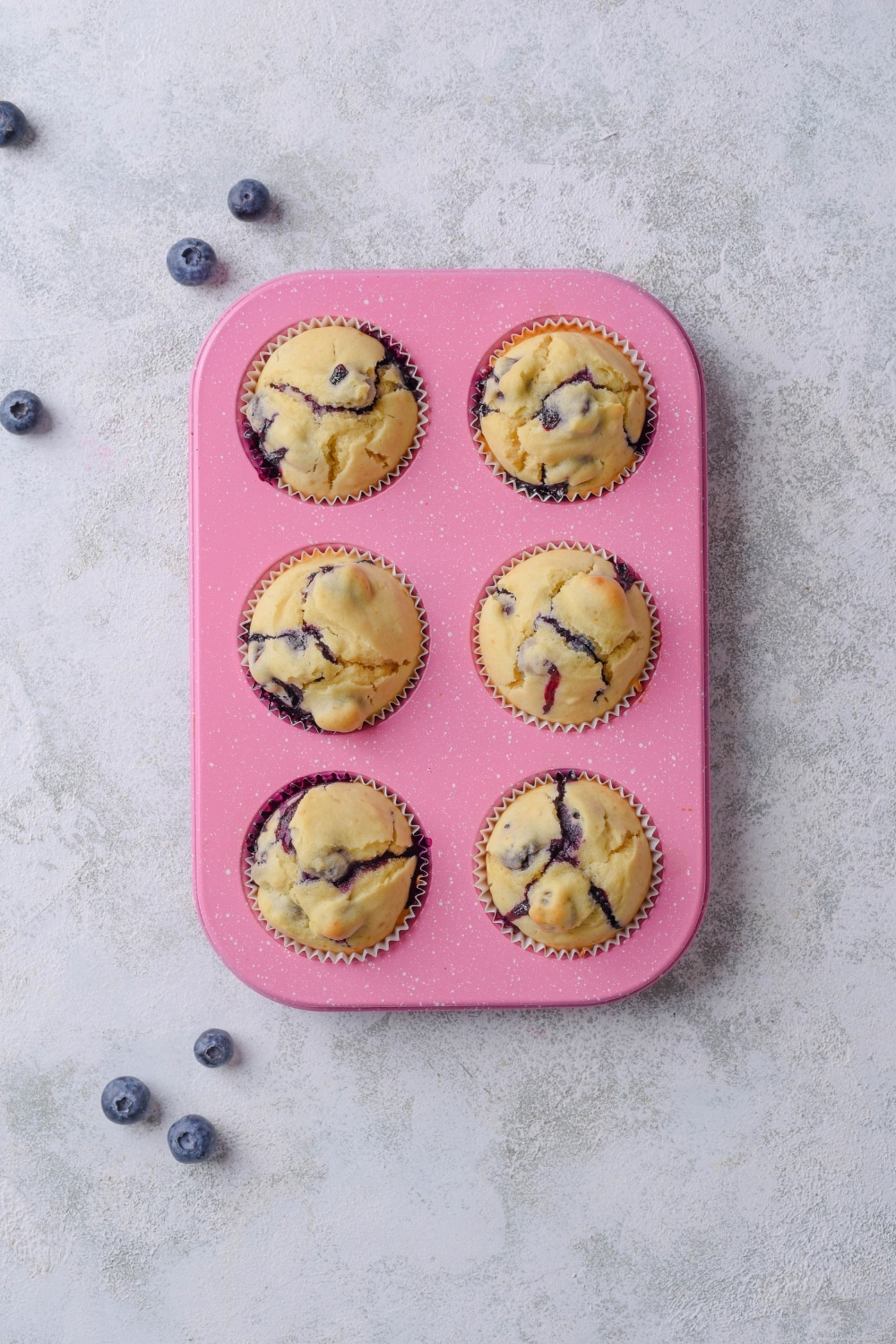 A pink muffin tray of six blueberry muffins fully baked. The tray sits on top of a marble counter.