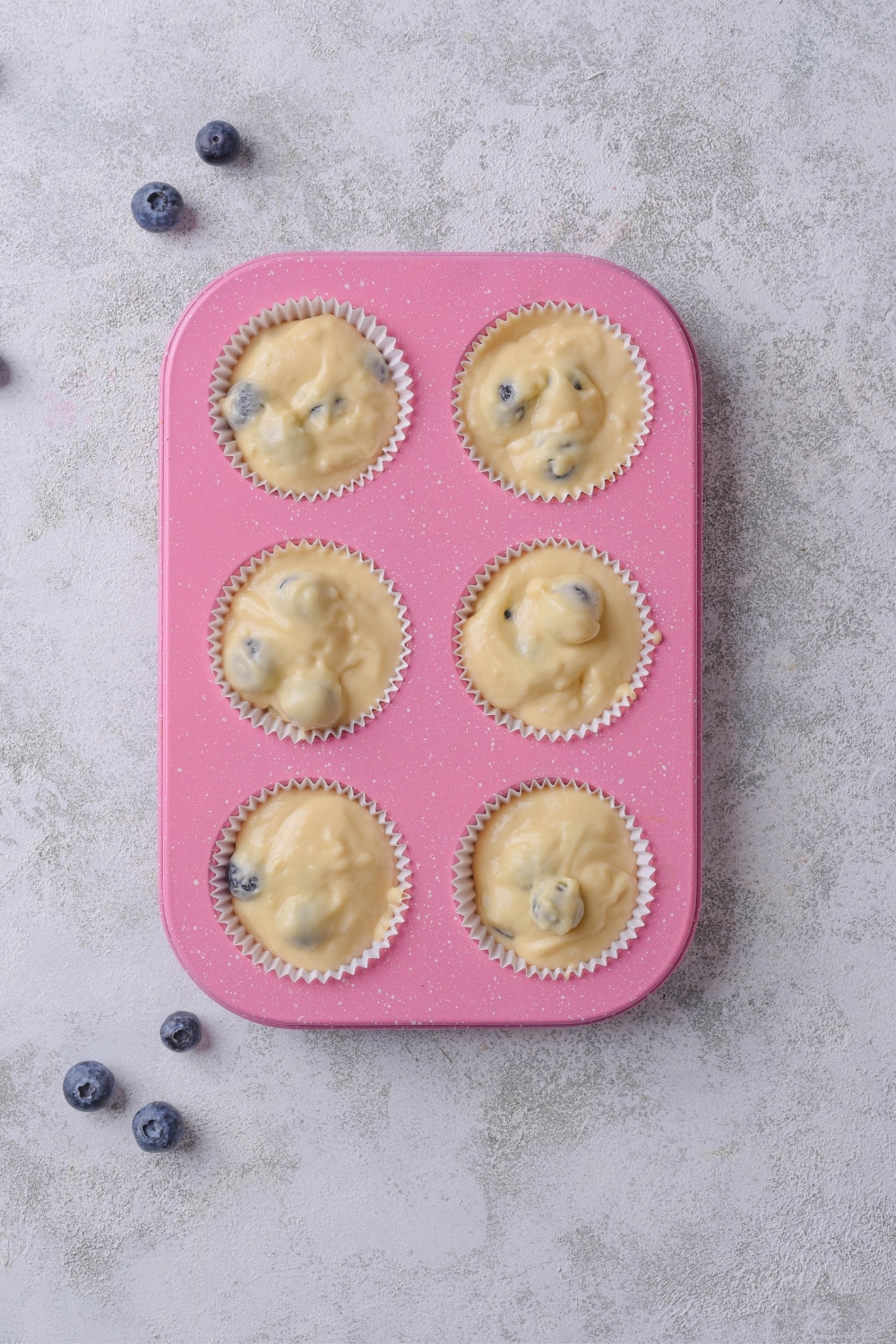 A pink muffin pan with six muffin liners filled with batter. The pan in on of a marble countertop.