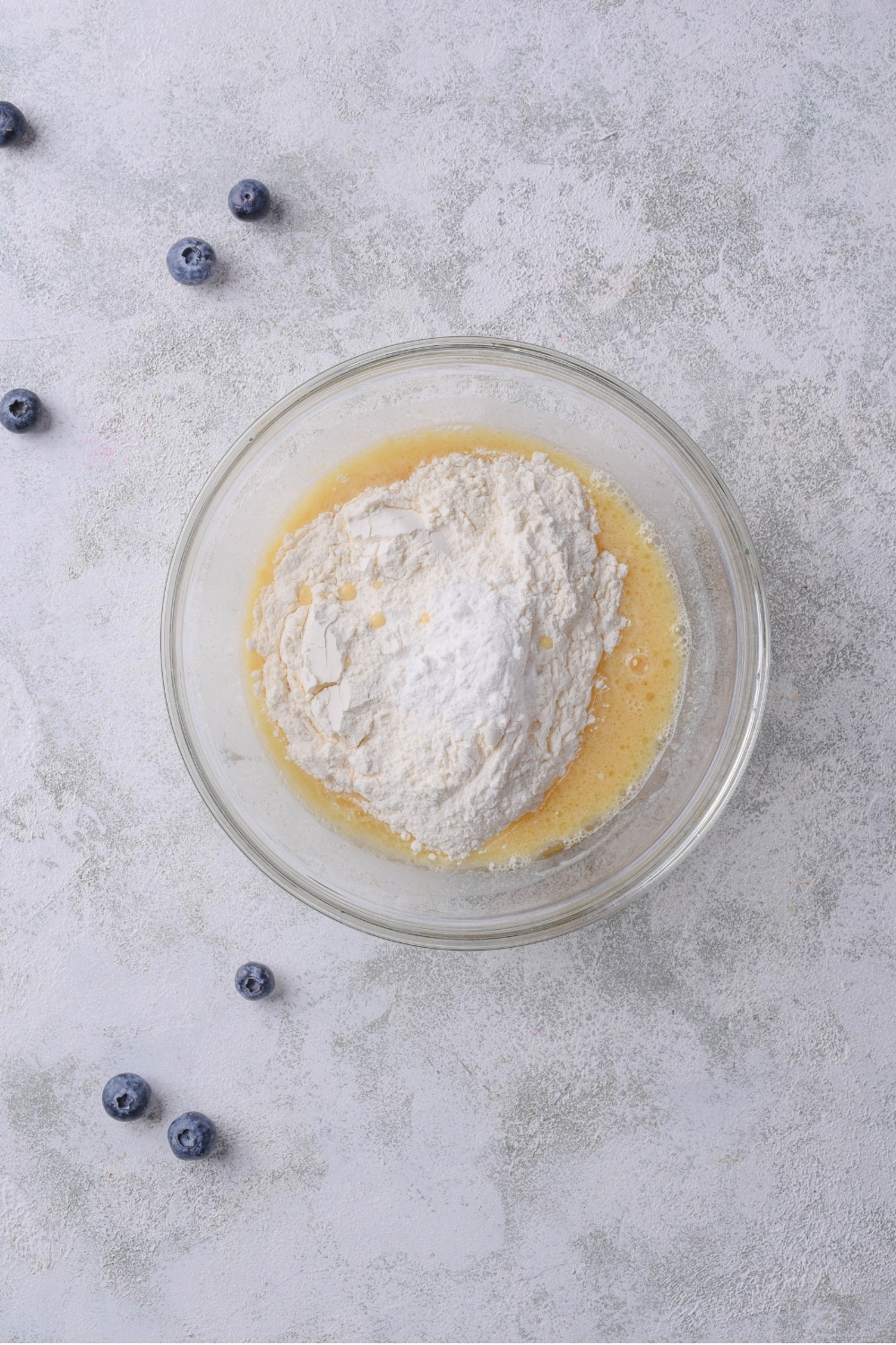 A glass bowl with an egg mixture and flour piled on top. The bowl sits on a marble countertop.