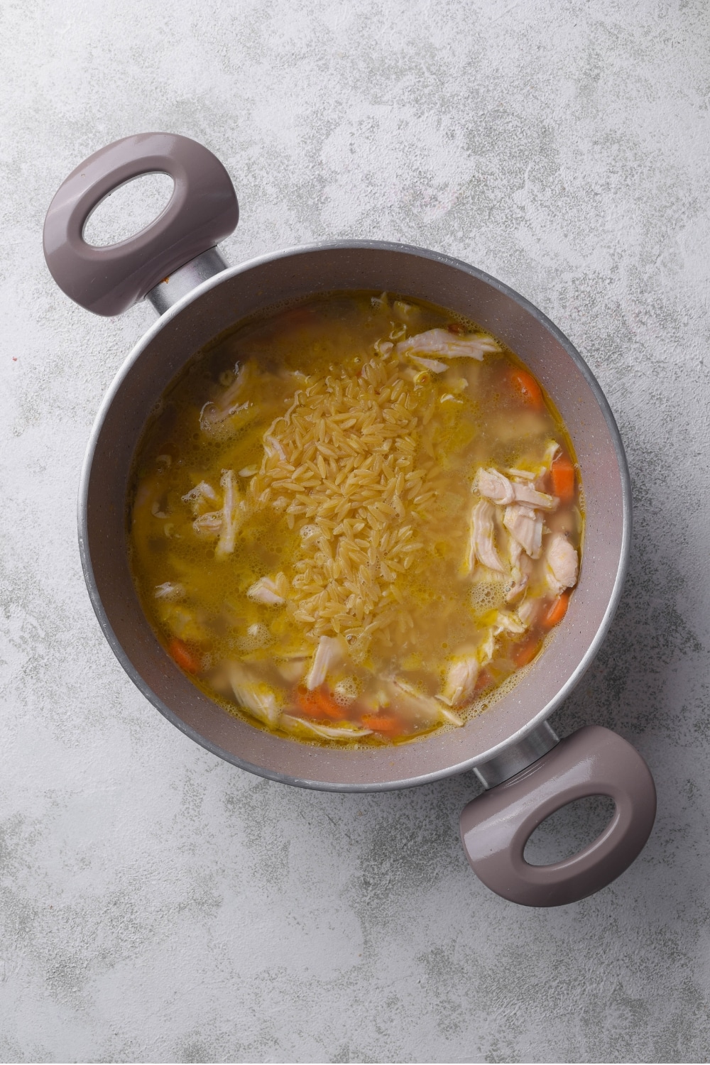 A large pot with shredded chicken, carrots, and orzo pasta cooking in chicken broth.