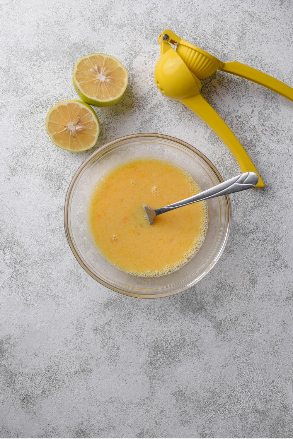 A small glass bowl with a mixture of whisked eggs, lemon juice, and broth. A lemon juicer and two lemon halves are next to the bowl.