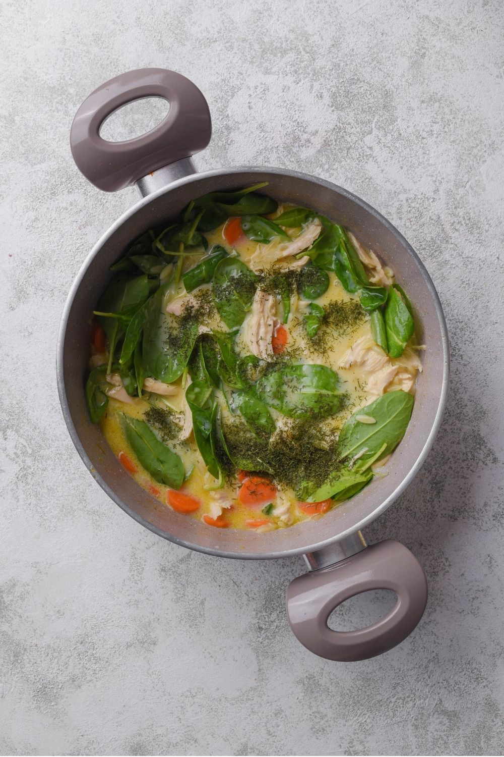 A large pot with creamy chicken broth, spinach, carrots, shredded chicken, and orzo pasta with seasonings sprinkled on top.