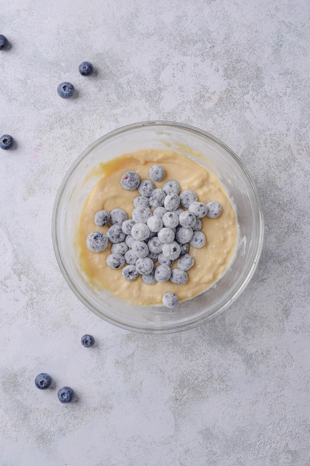 A glass bowl with blueberries piled on top of muffin batter. The bowl sits on a marble countertop.