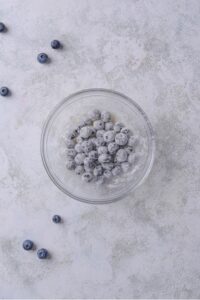 A glass bowl with blueberries dusted with flour on a marble countertop.