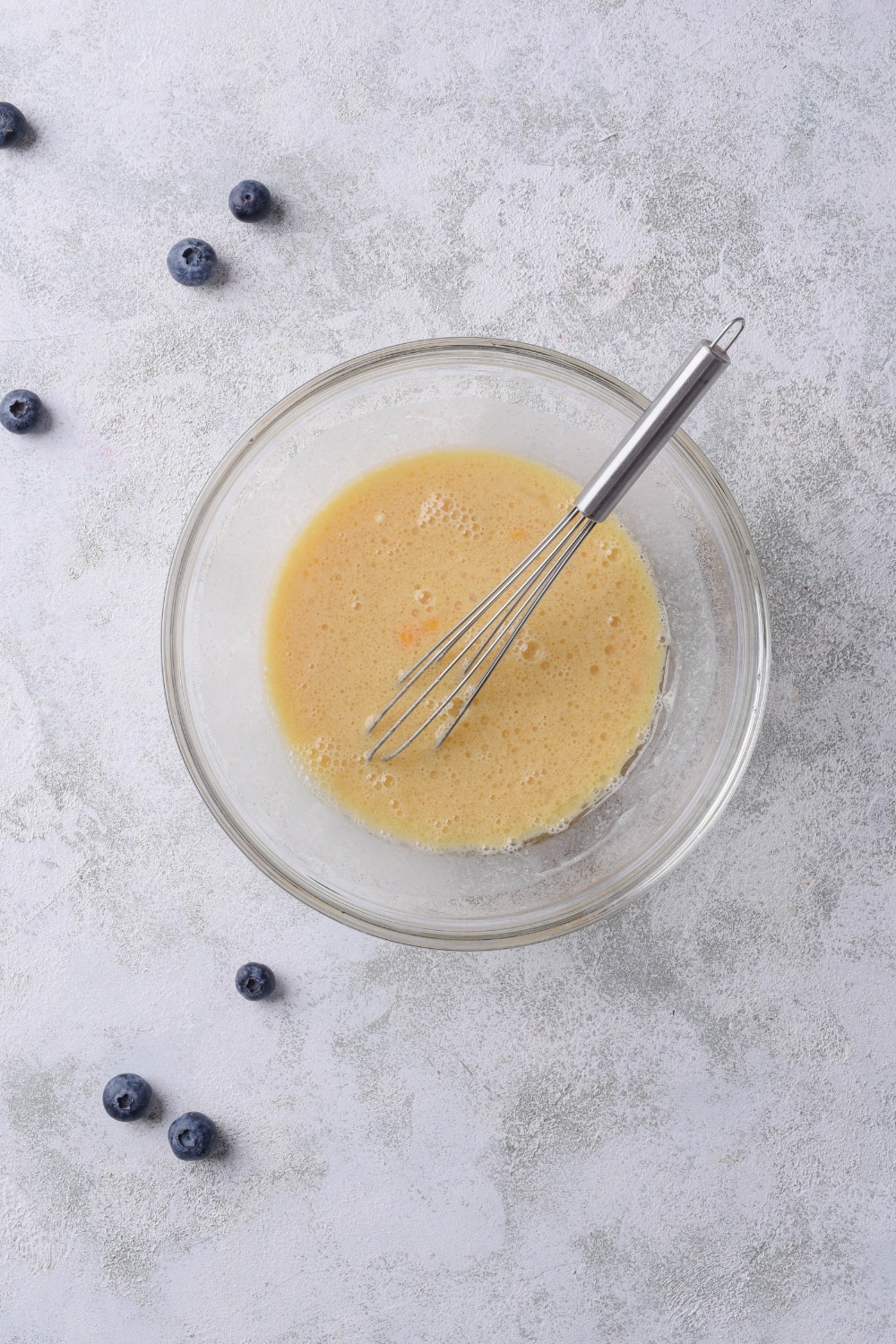 A glass bowl with a whisk submerged in an egg mixture on a marble countertop.