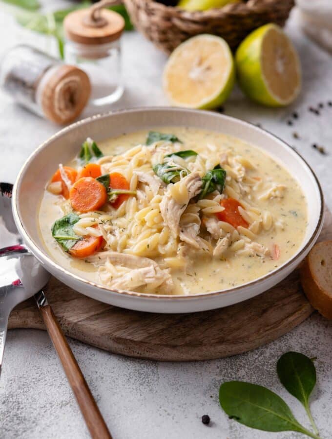 A white ceramic bowl of lemon chicken soup with orzo pasta, carrots, and spinach. The bowl sits on a wooden board with two spoons on the side.