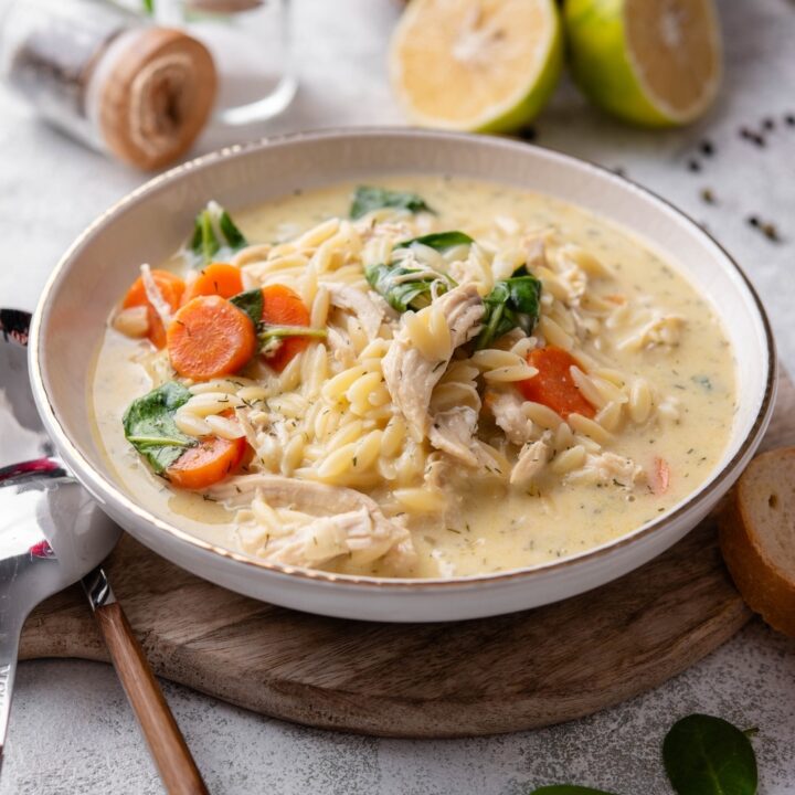 A white ceramic bowl of lemon chicken soup with orzo pasta, carrots, and spinach. The bowl sits on a wooden board with two spoons on the side.