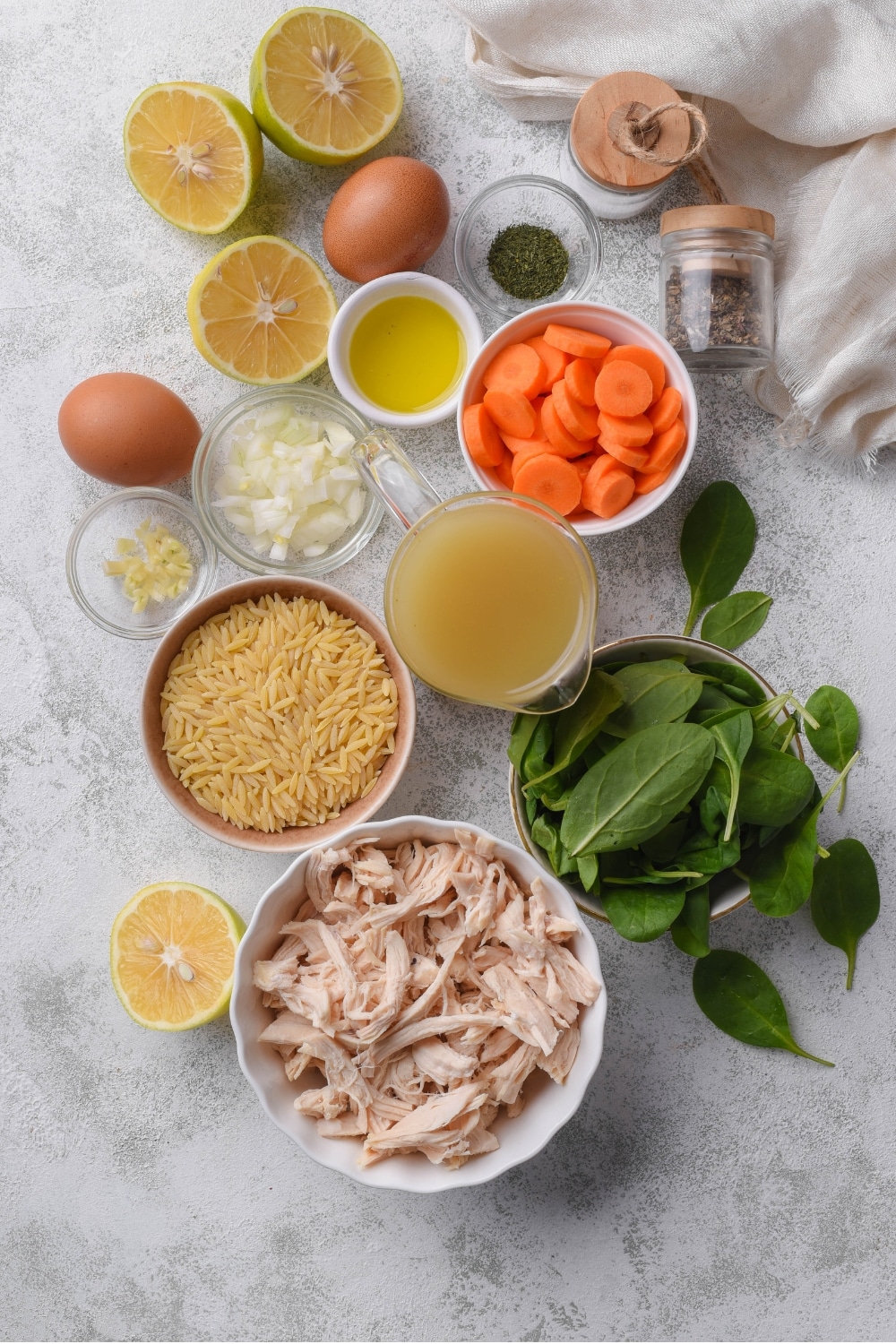 Several bowls of varying soup ingredients including shredded chicken, spinach, orzo pasta, broth, carrots, garlic, onion, and herbs.