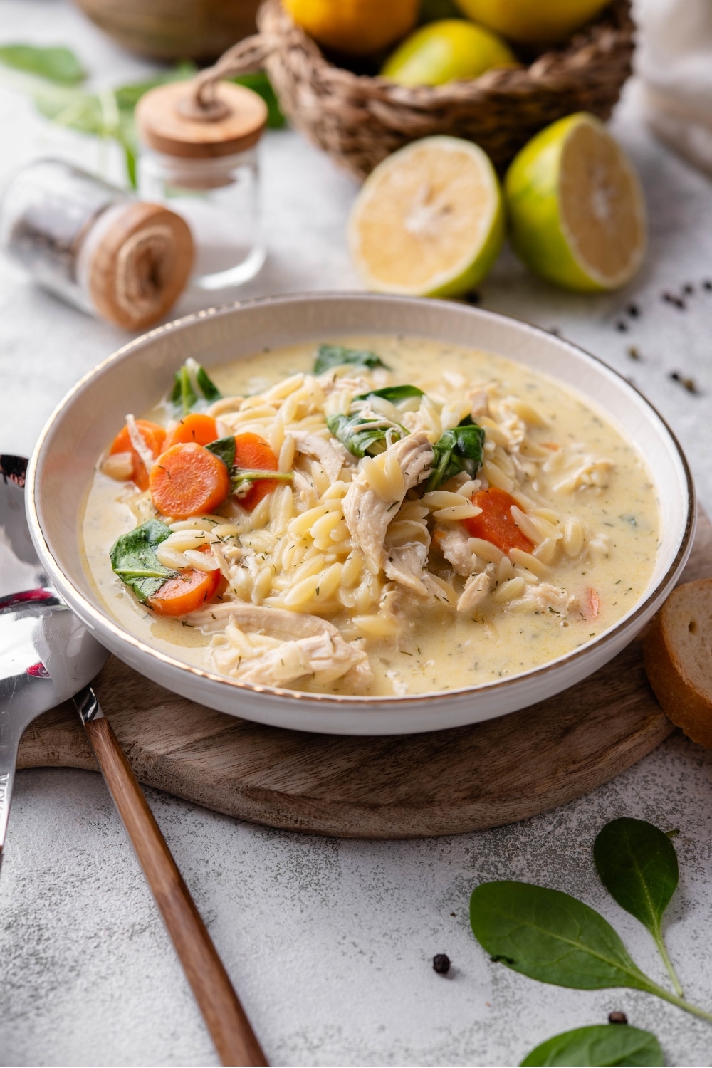 A white ceramic bowl of lemon chicken soup with orzo pasta, carrots, and spinach. The bowl sits on a wooden board with two spoons on the side.
