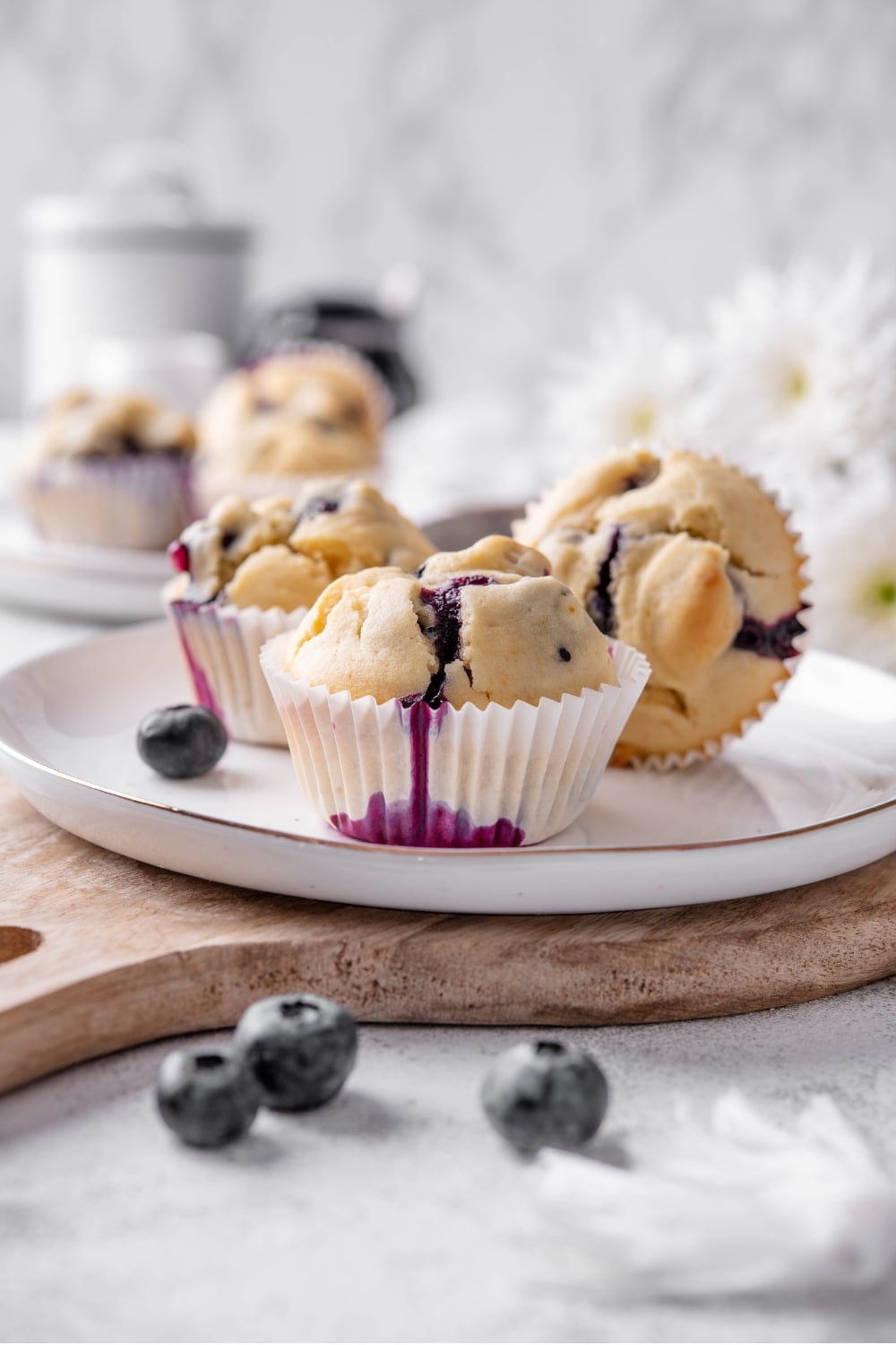 Three blueberry muffins sit on a white ceramic plate atop a wooden serving board. There is an extra plate of muffins and white flowers in the background