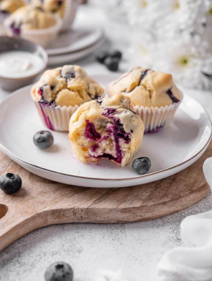 Three blueberry muffins sit on a white ceramic plate atop a wooden serving board. There is a bite taken out of a muffin with an extra plate of muffins and white flowers in the background