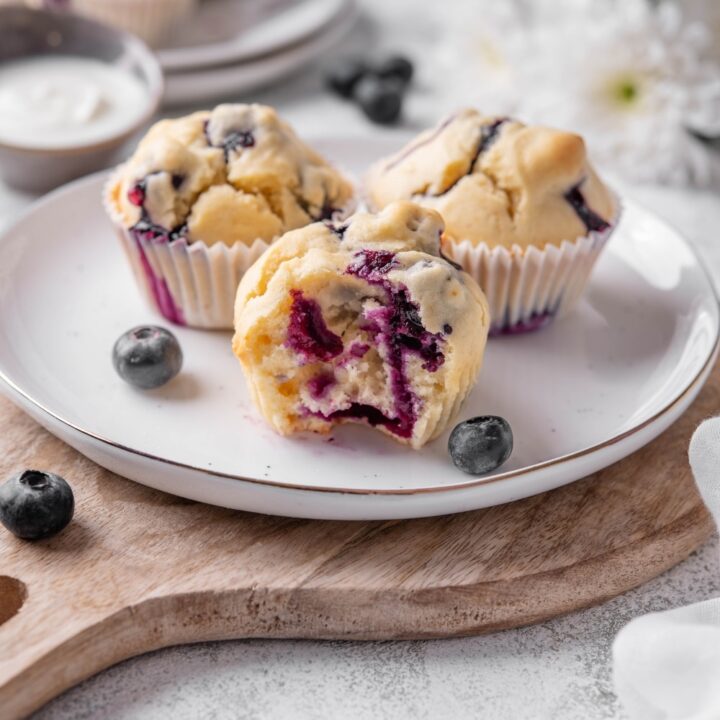 Three blueberry muffins sit on a white ceramic plate atop a wooden serving board. There is a bite taken out of a muffin with an extra plate of muffins and white flowers in the background