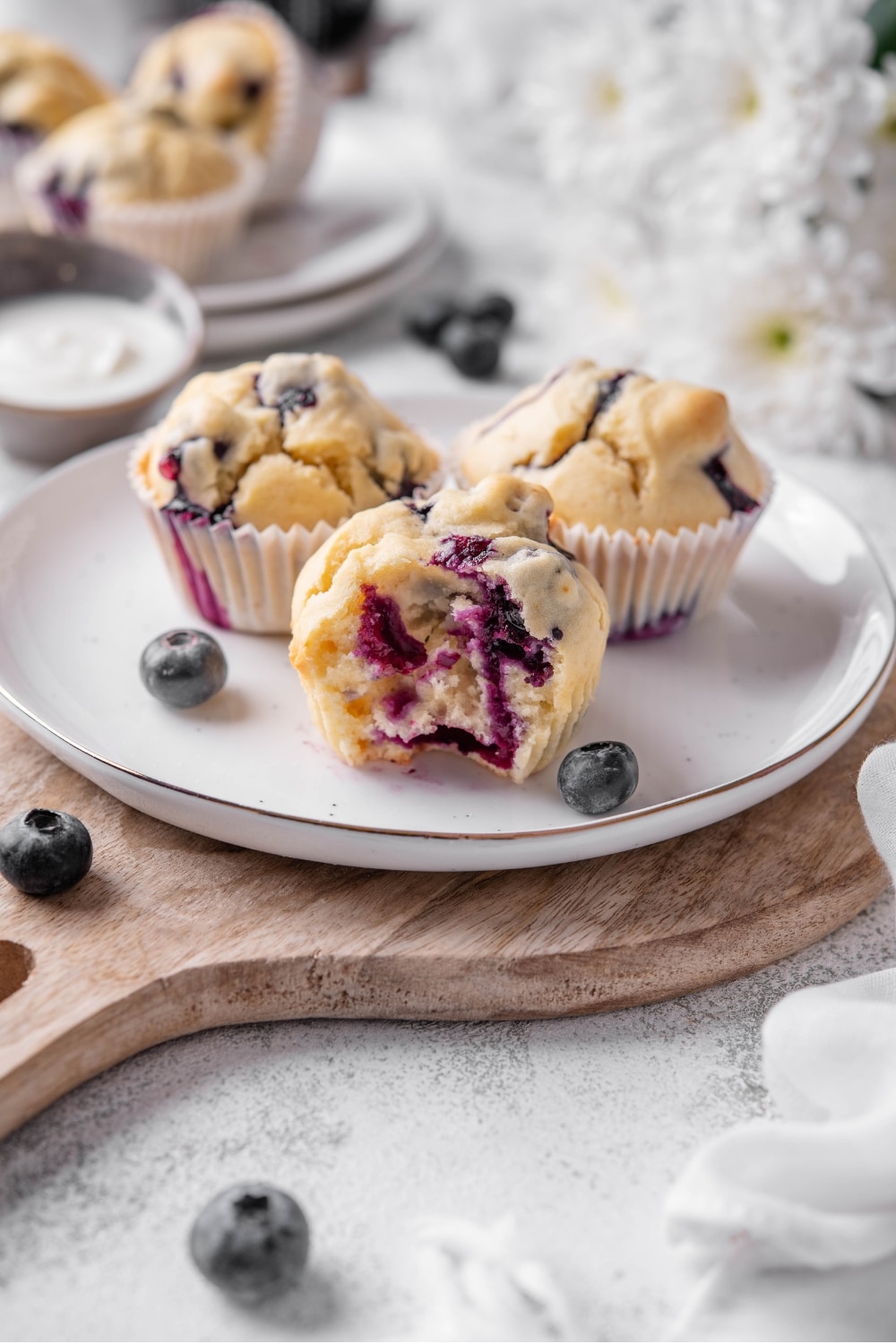 Three blueberry muffins sit on a white ceramic plate atop a wooden serving board. There is a bite taken out of a muffin with an extra plate of muffins and white flowers in the background