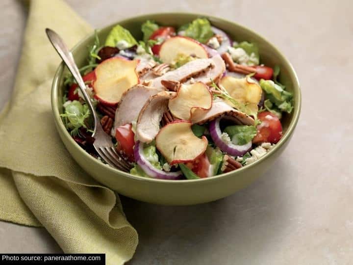 A few apple crisps, red onion, tomato, and sliced chicken in a green bowl with a fork in it.