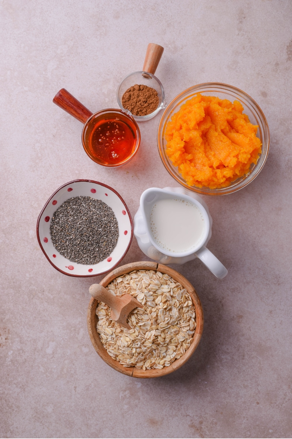 Several bowls of varying designs and sizes carrying ingredients such as pumpkin, oats, milk, chia seeds, honey, and pumpkin pie spice.