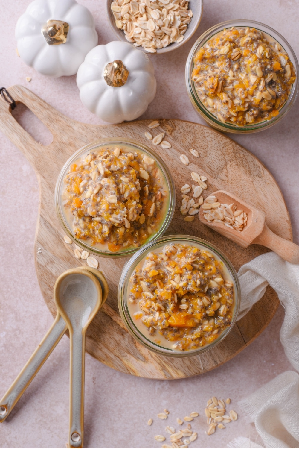 Two jars of pumpkin overnight oats sit on a wooden board with two ceramic measuring spoons next to them. There is a third jar of overnight oats next to the board along with two white pumpkins.