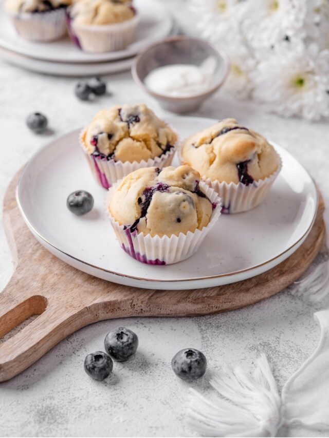 Three blueberry muffins sit on a white ceramic plate atop a wooden serving board. There is an extra plate of muffins and white flowers in the background
