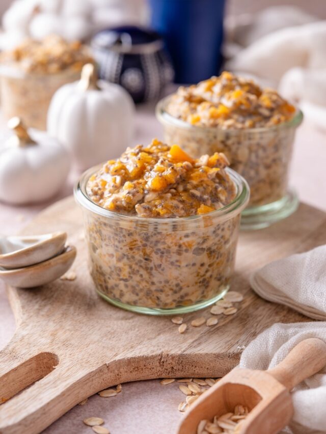 Two glass jars filled with pumpkin overnight oats on a wooden serving board. There are decorative white pumpkins and ceramic spoons next to them.