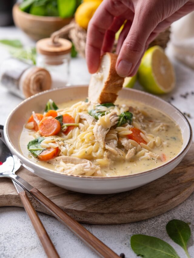 A white ceramic bowl of lemon chicken soup with orzo pasta, shredded chicken, carrots, and spinach. A hand is dipping a slice of bread into the soup.