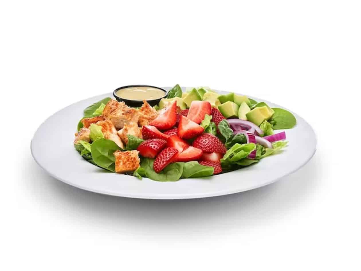 Sliced strawberries, chicken, avocado, red onion, baby spinach, and cup of dressing on a white plate.