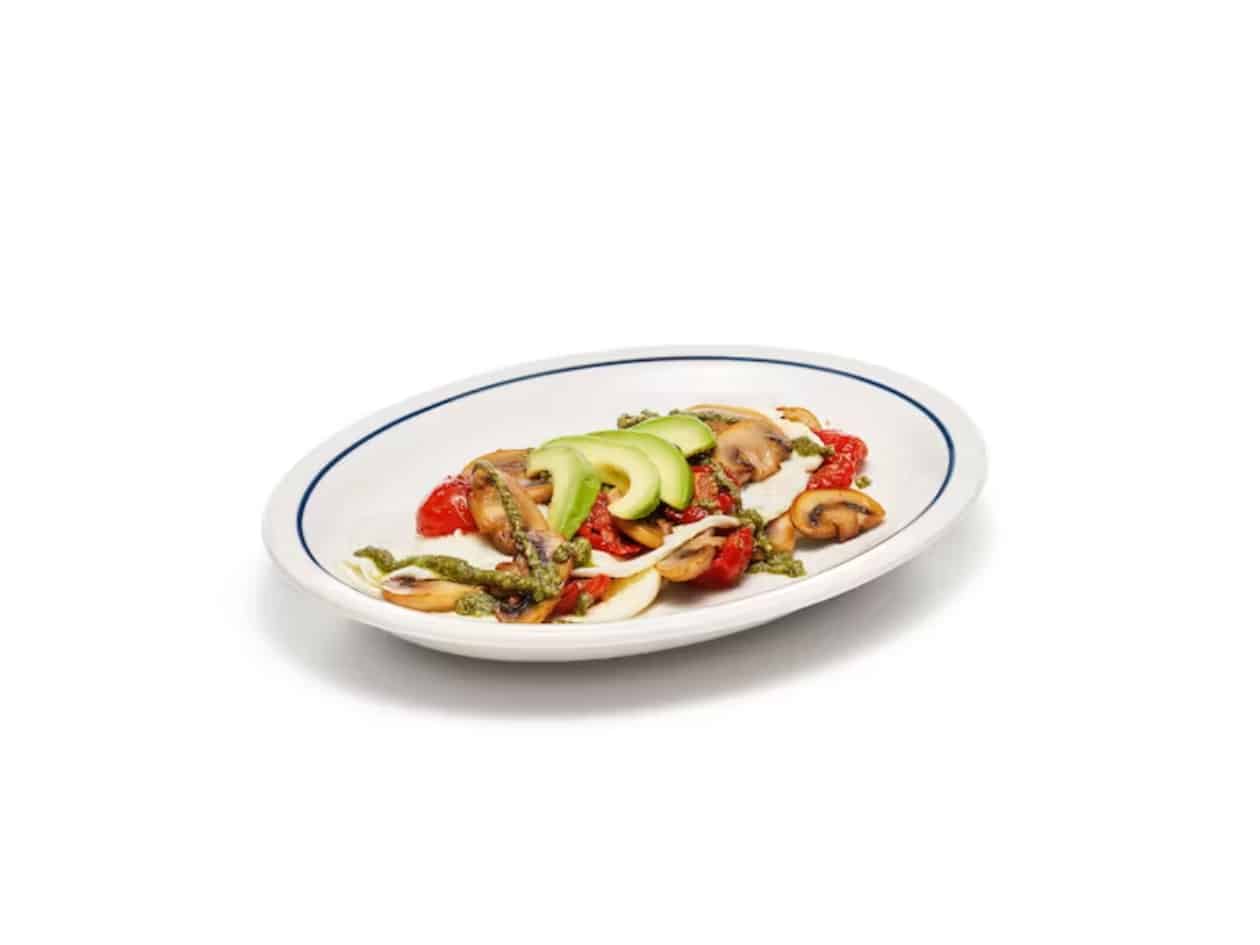 Sliced avocado, mushrooms, tomatoes, and pesto on egg whites on a white plate.